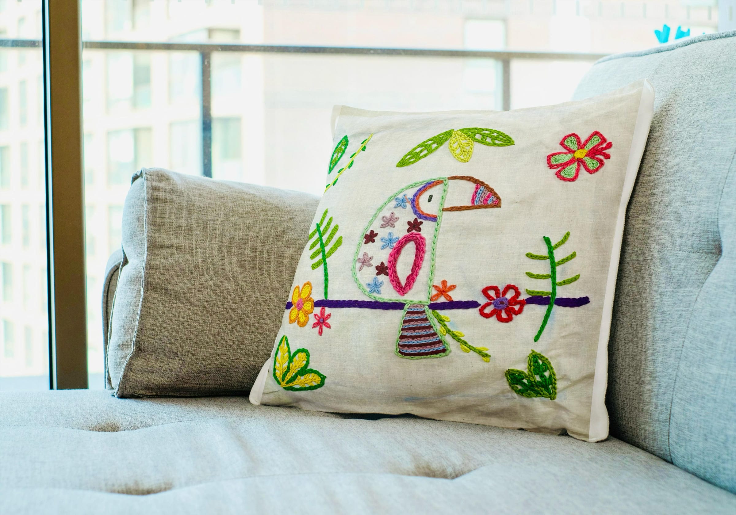 White cushion embroidered with a colourful toucan and flowers on a grey sofa