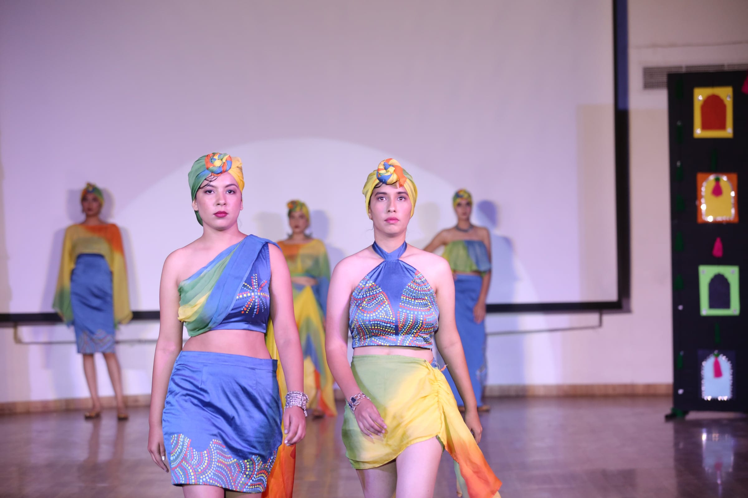 Two models in teal and saffron looks walking the runway towards the camera